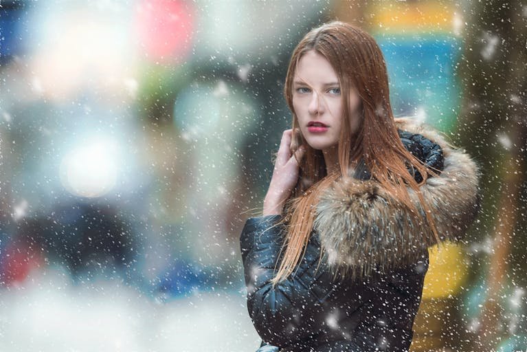 A fashionable woman in a fur coat poses elegantly amidst falling snow in a vibrant cityscape.