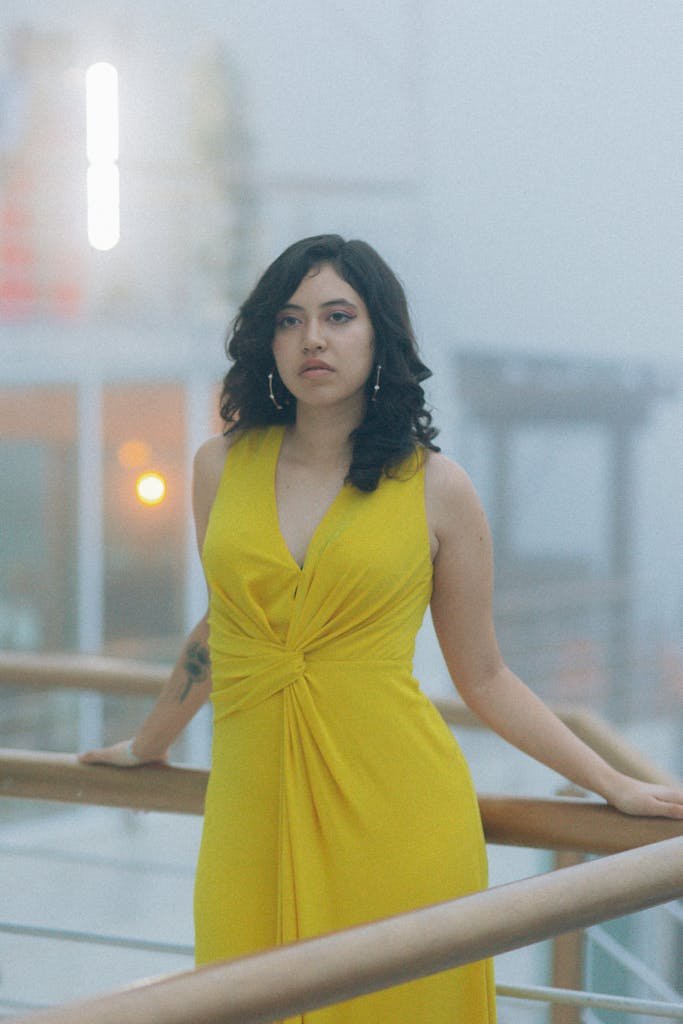 Stylish woman in a yellow dress poses calmly on a foggy outdoor balcony, exuding elegance.